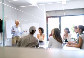 a person holding the attention of seven office workers inside a bright meeting room